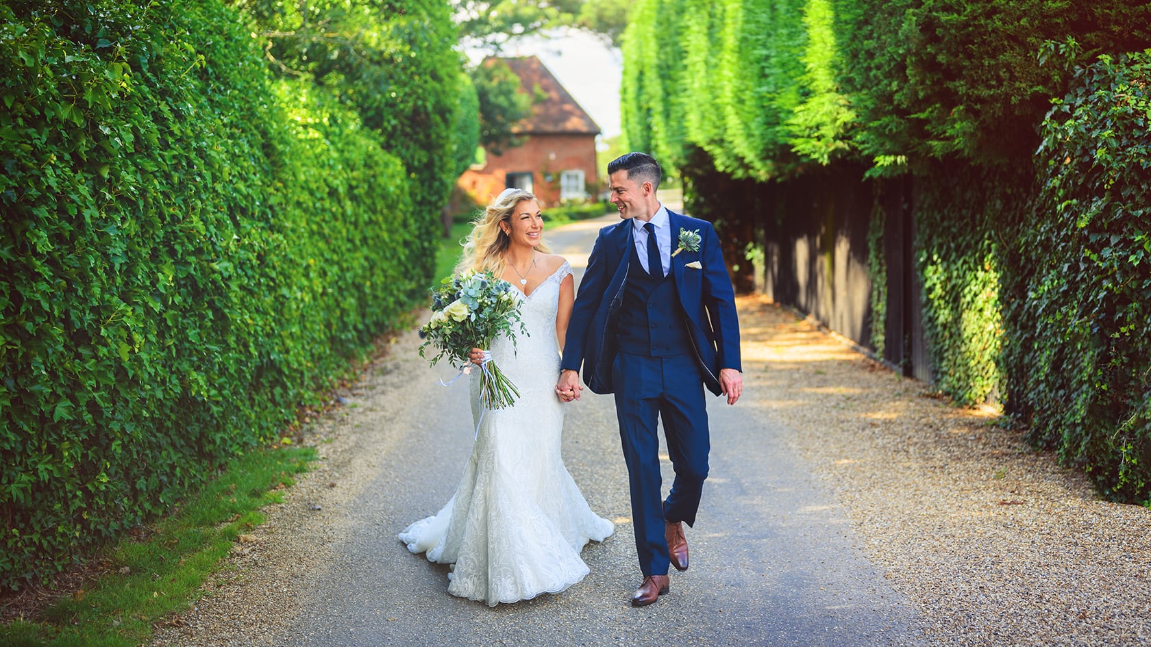 A newly married couple walking along the drive at Gaynes Park, with a cottage behind them.